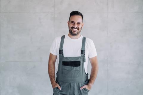 Ein lächelnder Mann in grauer Latzhose und weißem T-Shirt steht vor einer hellgrauen Betonwand und hat die Hände in den Hosentaschen.
