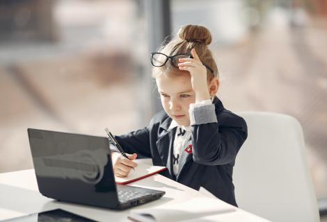 Ein kleines Mädchen in Businesskleidung sitzt konzentriert an einem Schreibtisch mit Laptop und Notizbuch. Sie hebt ihre Brille an die Stirn, hält einen Stift in der Hand und wirkt fokussiert, als ob sie arbeitet oder etwas Wichtiges plant.
